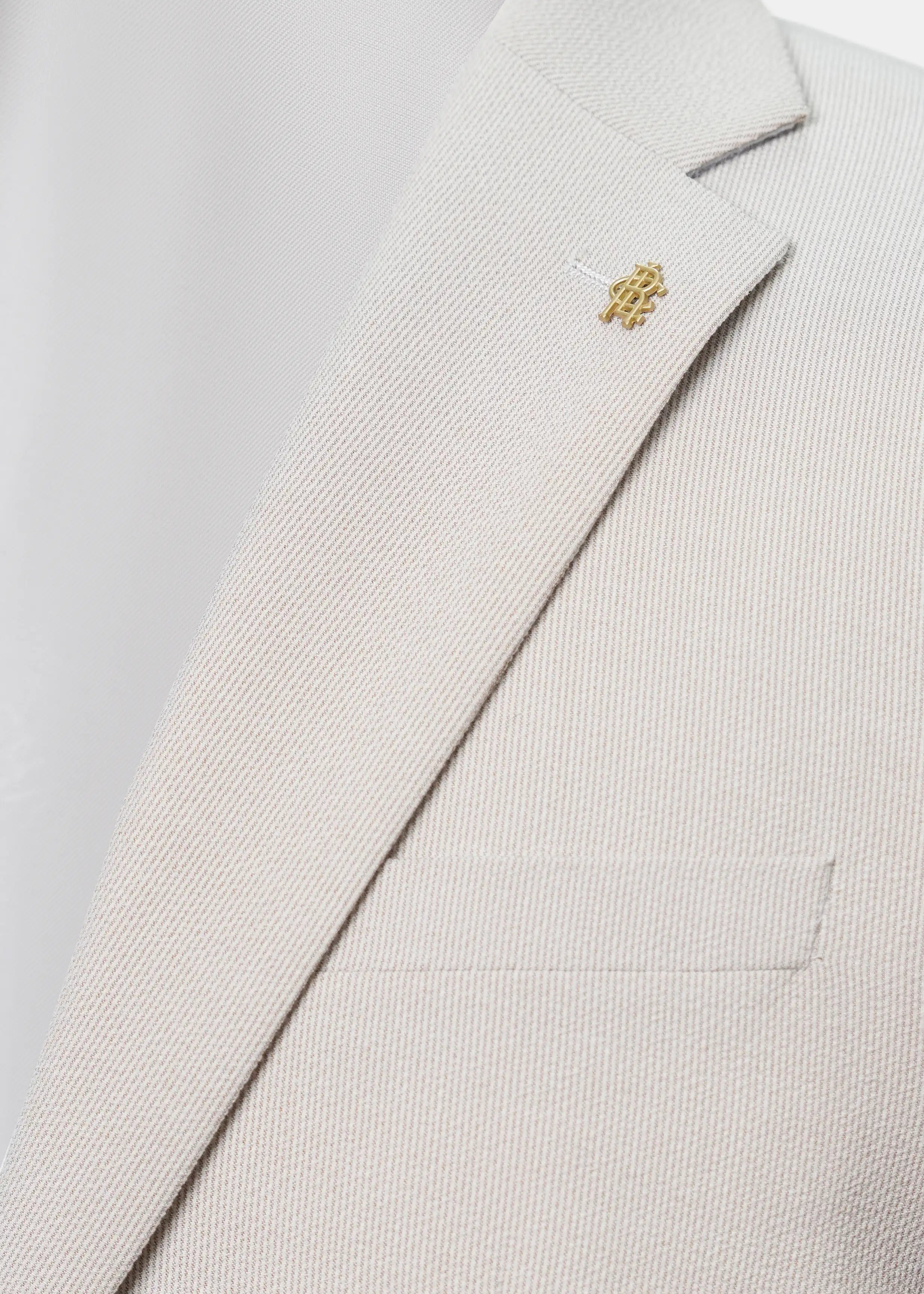 Beige casual coat lapel detail with small gold emblem pin